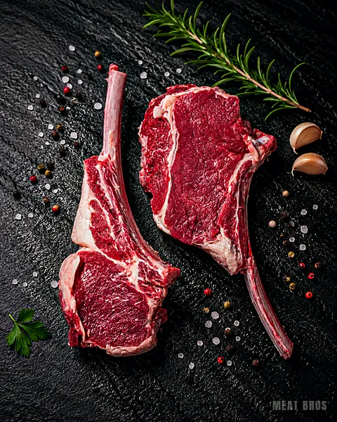 Dos tomahawk de res crudos con hueso largo sobre piedra negra, con romero y ajo