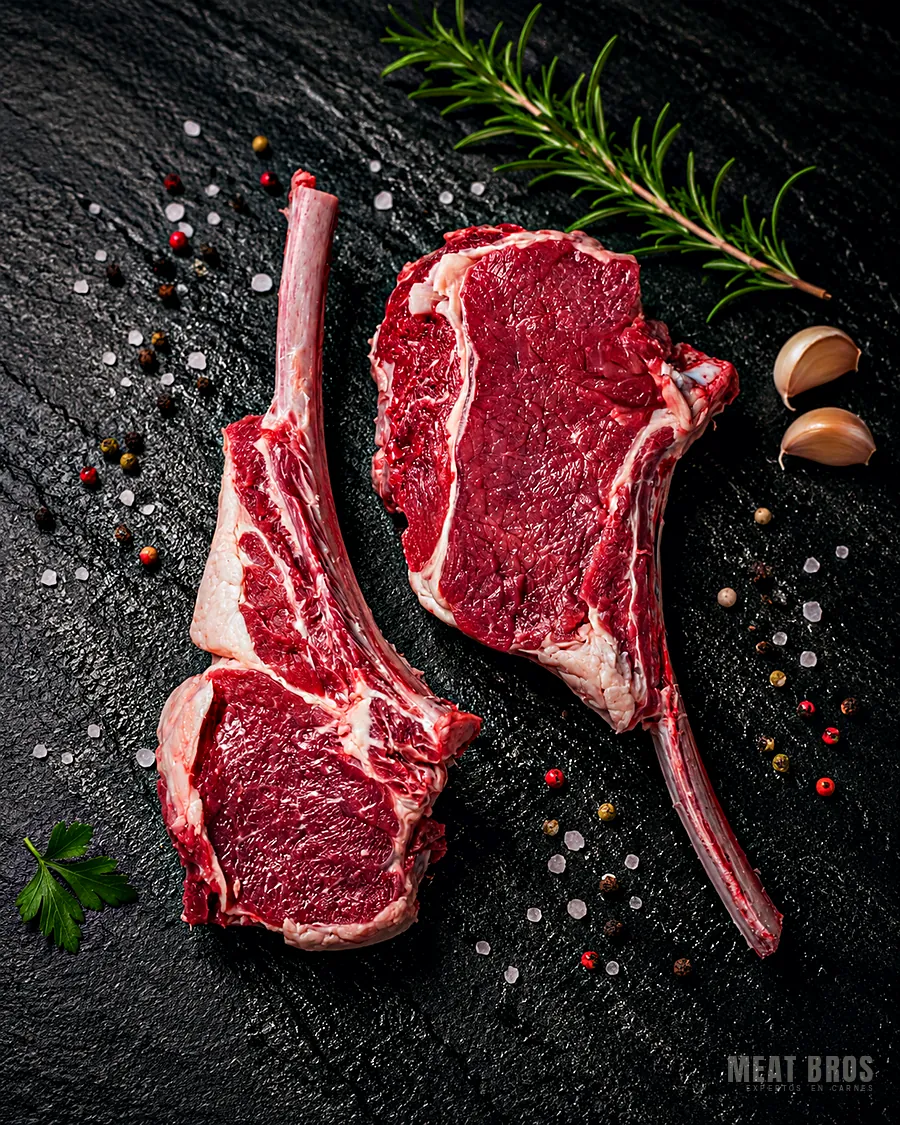 Dos tomahawk de res crudos con hueso largo sobre piedra negra, con romero y ajo